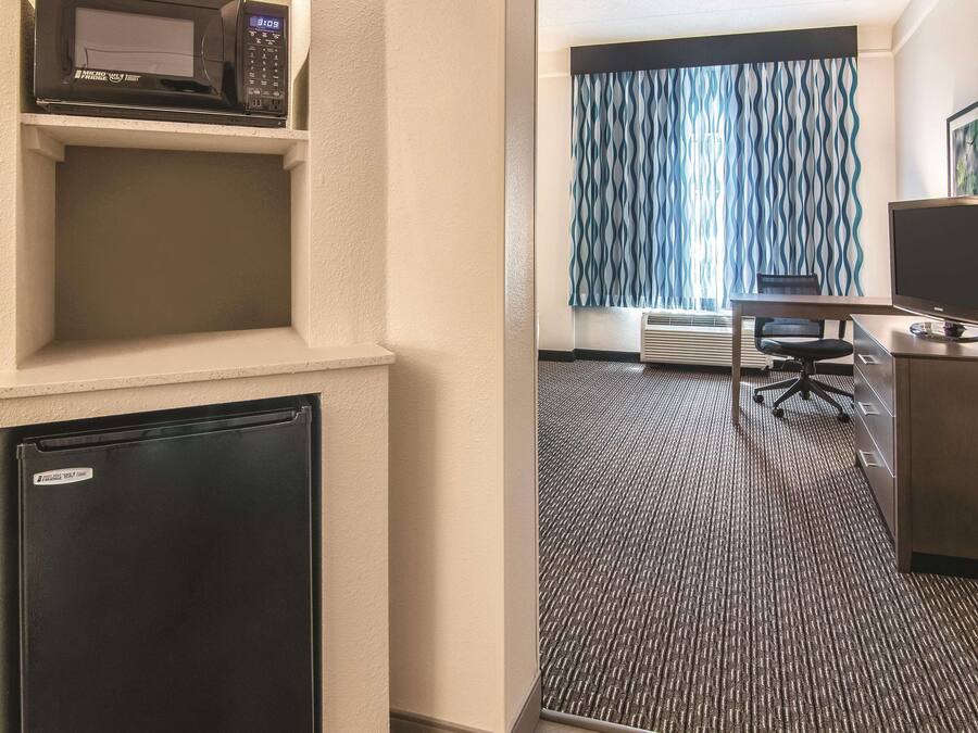 A hotel room with a microwave, a small refrigerator, a desk with a computer, a chair, and a patterned curtain.