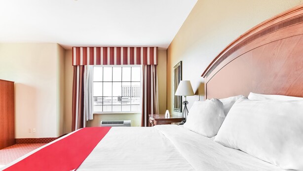 A hotel room with a large bed, a red and white striped curtain, a window with a view of buildings, a bedside table with a lamp, and a wooden headboard.