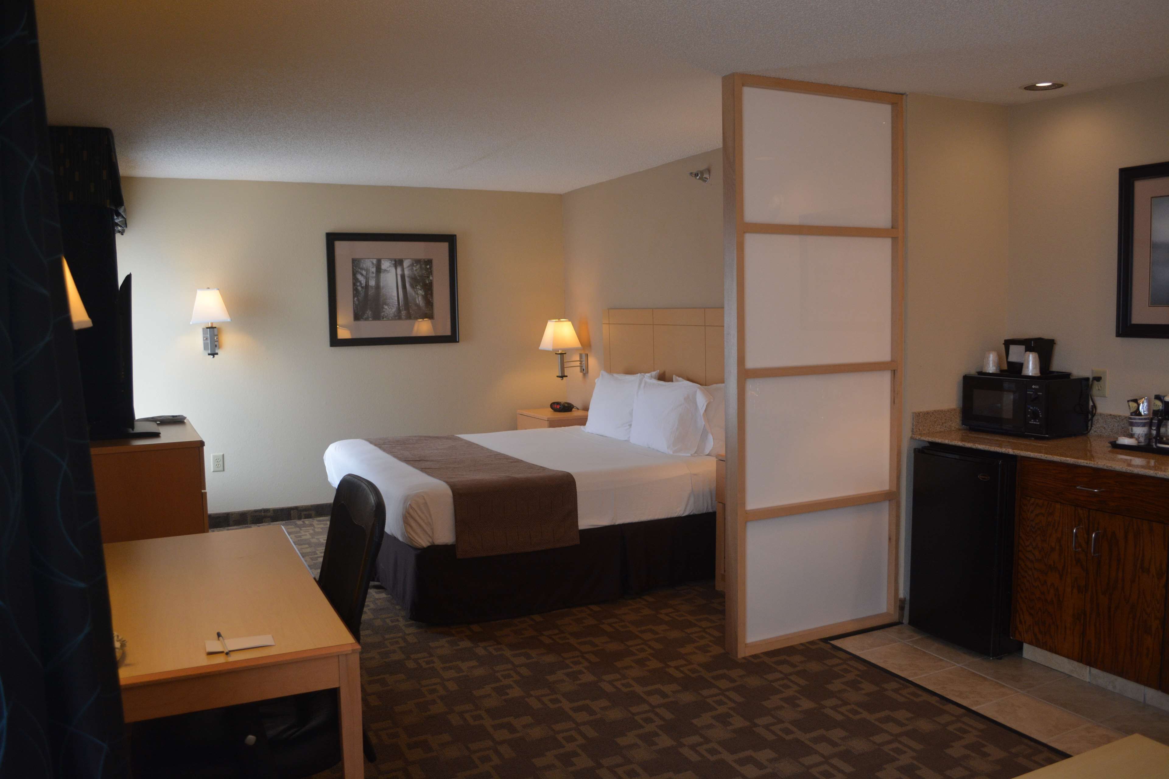 A hotel room with a bed, bedside table, lamp, and a small kitchenette area.