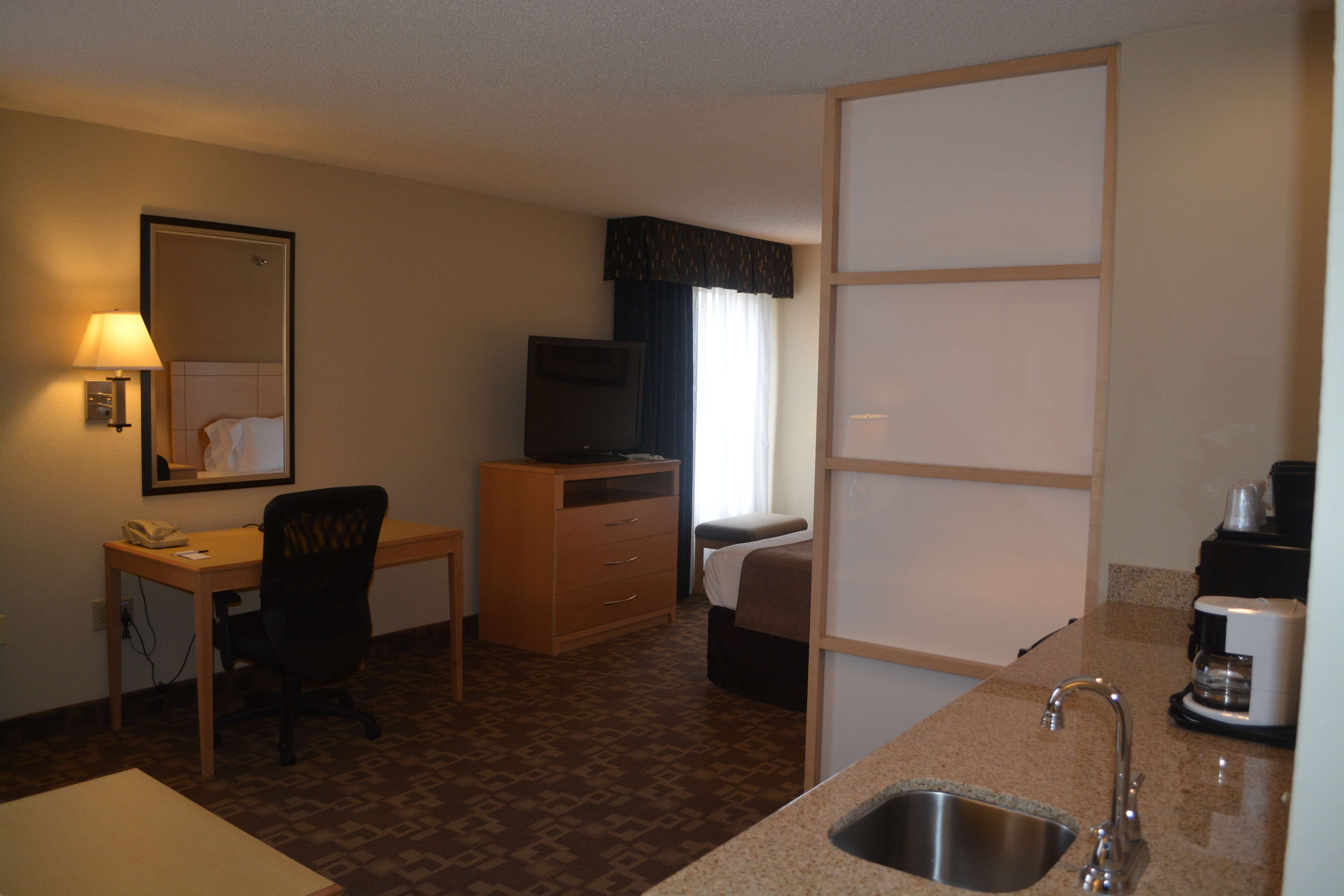 Una habitación de hotel compacta con kitchenette, escritorio, silla, cama, televisor y ventana con cortinas.