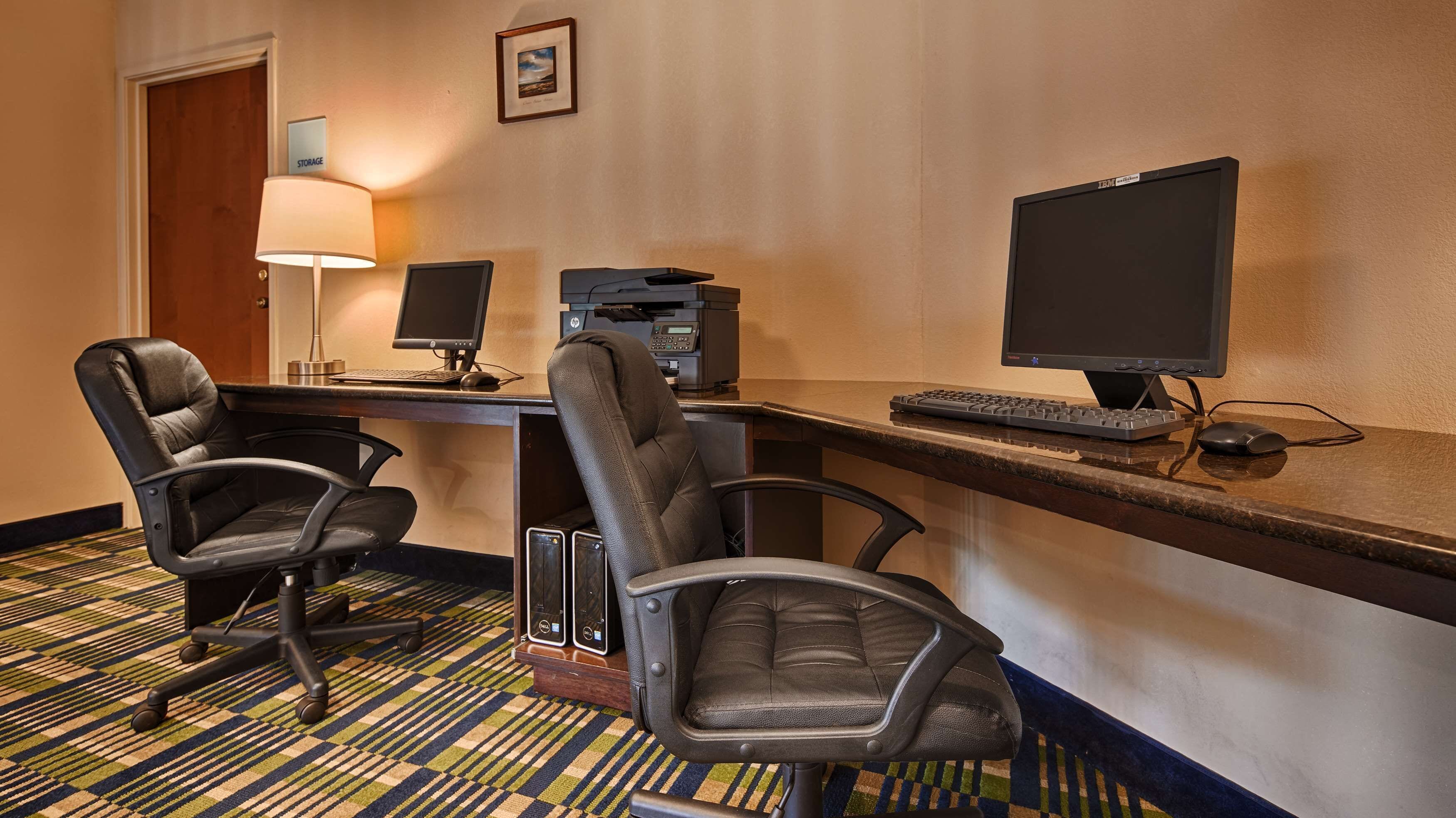 A hotel room with a desk, two chairs, a computer, a printer, and a lamp.