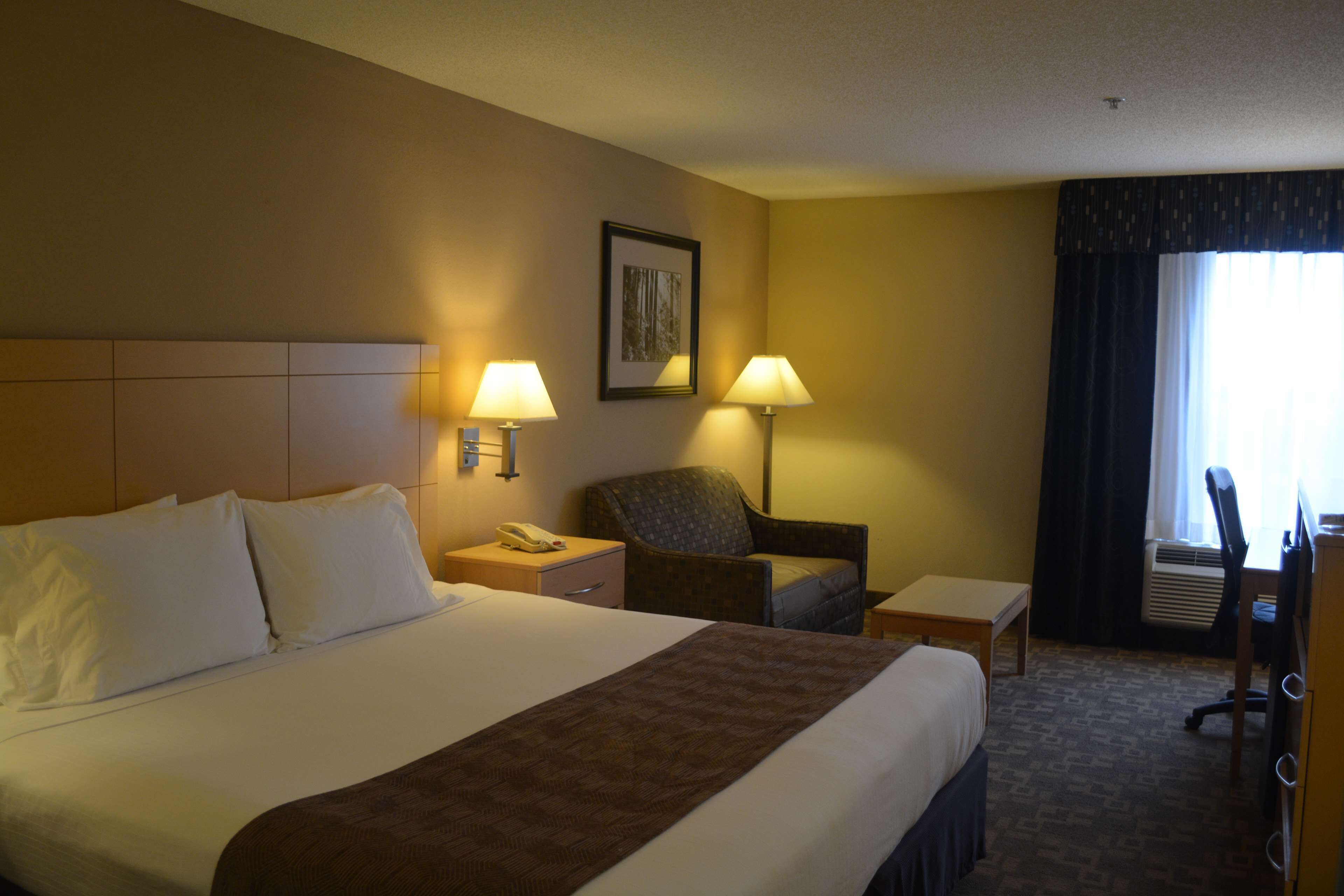A hotel room with a large bed, two bedside lamps, a chair, a desk, and a window with curtains.