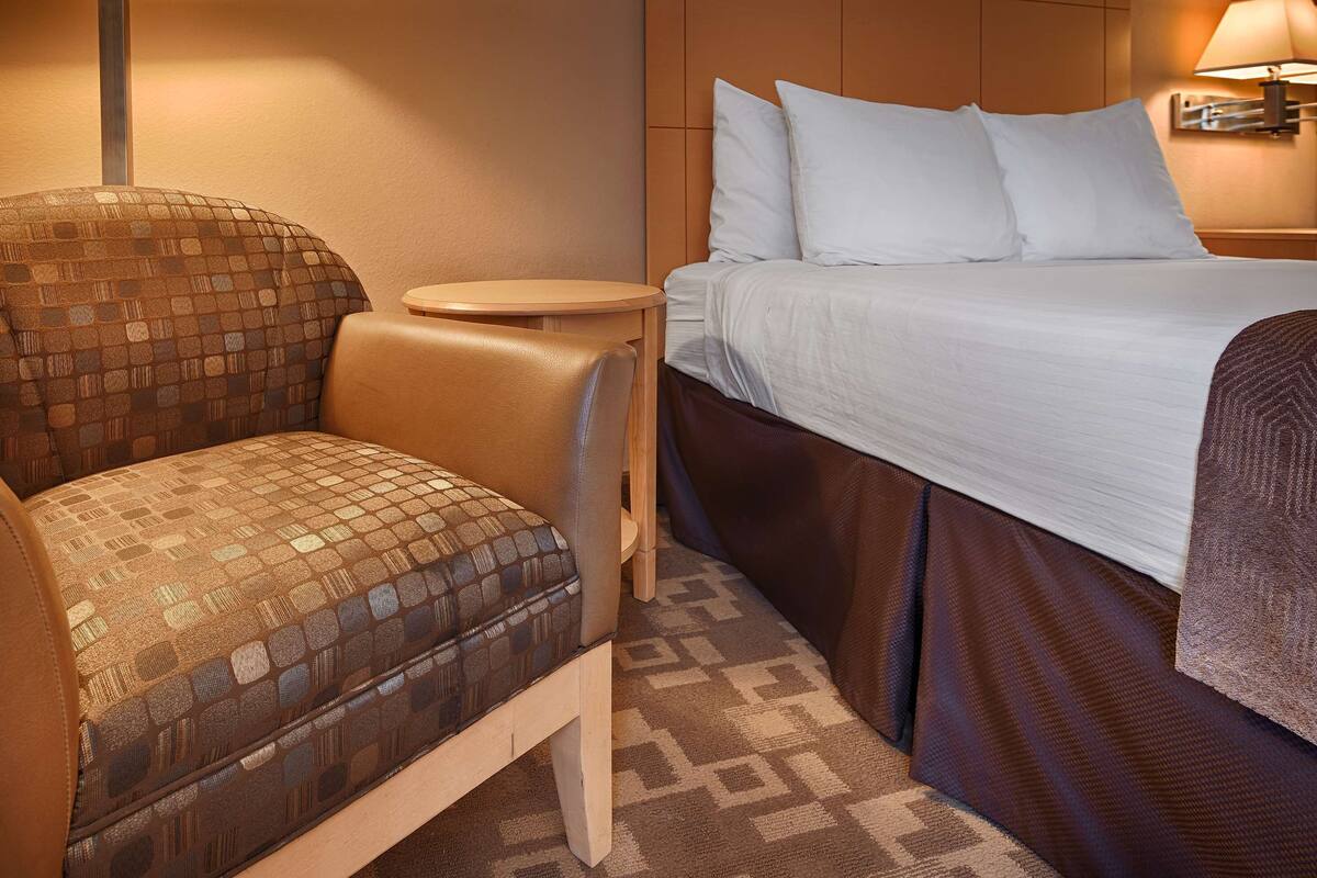 A hotel room with a bed, a chair, a small table, and a lamp.
