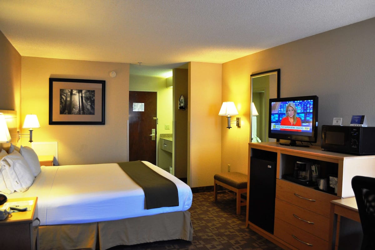 A hotel room with a bed, bedside lamps, a television displaying a news program, a microwave, a small table, and a chair.