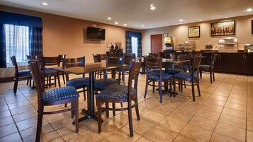 A hotel dining area with multiple tables and chairs, a kitchen area, and a television mounted on the wall.