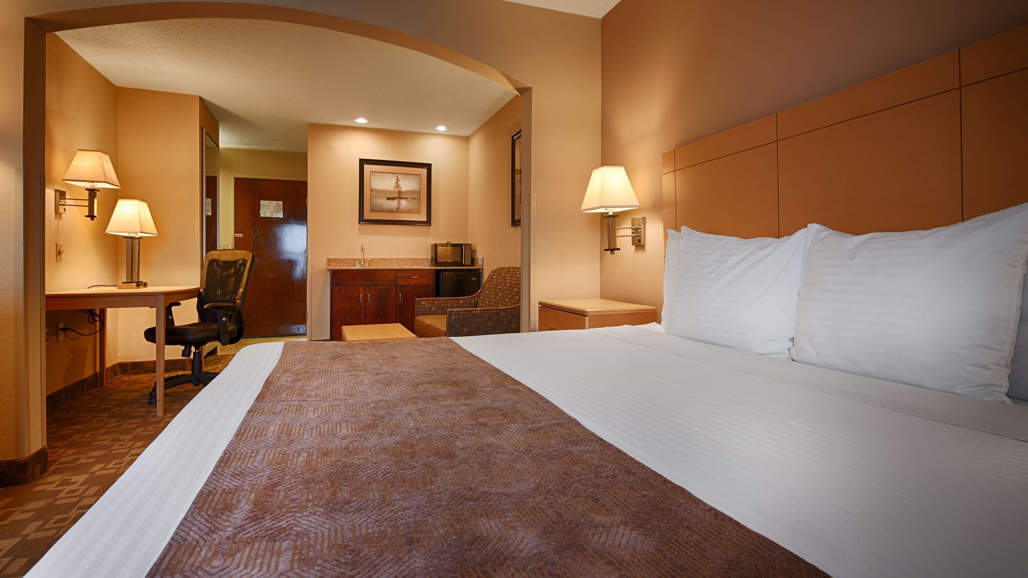 A hotel room with a large bed, a desk with a lamp, a chair, a small kitchenette, and a television.