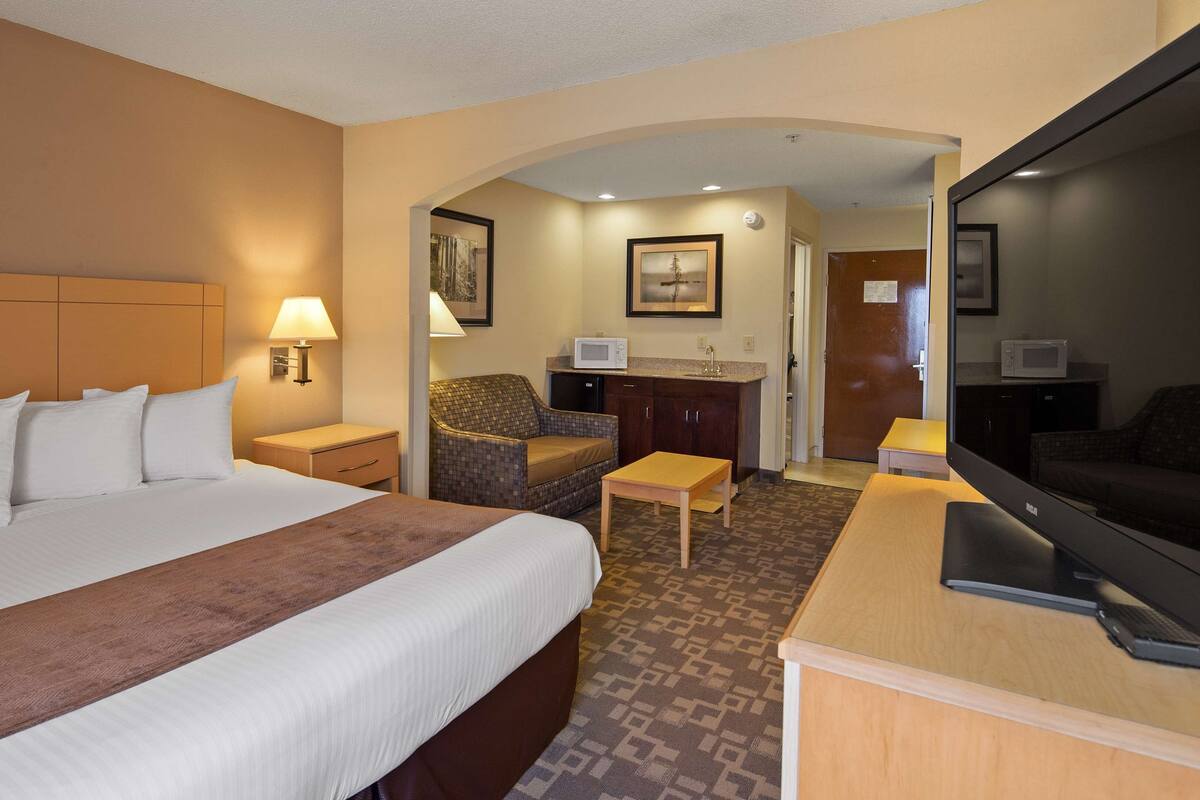 A hotel room with a large bed, a desk with a TV, a chair, a small table, and a kitchenette area.
