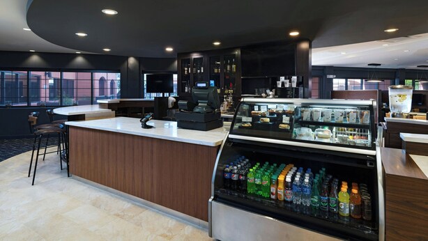 Un moderno vestíbulo de hotel con un mostrador de recepción curvo, una vitrina refrigerada para bebidas y un área de descanso con taburetes de bar.