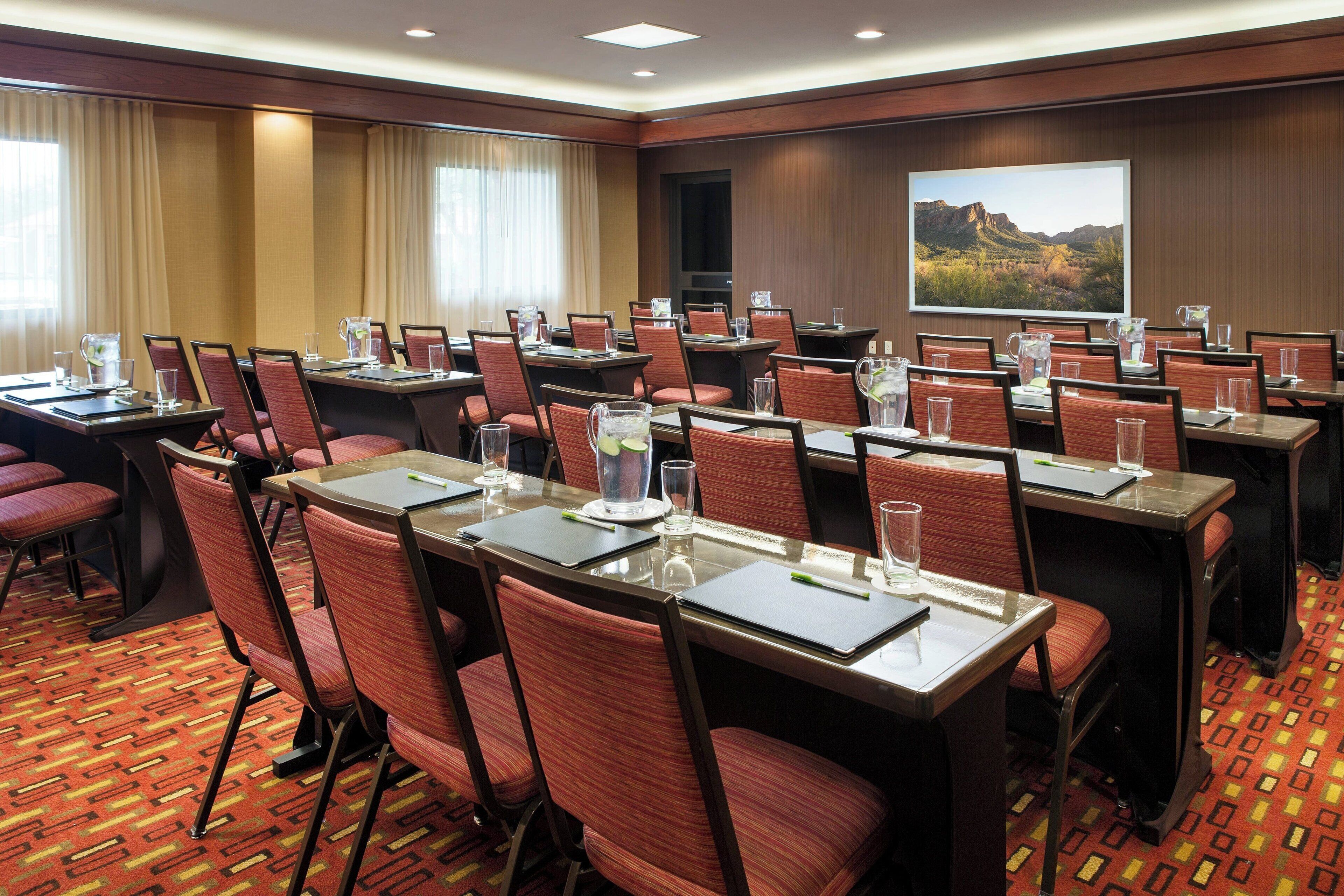 A conference room with multiple tables and chairs, equipped with water pitchers and glasses, and a large screen displaying a landscape.