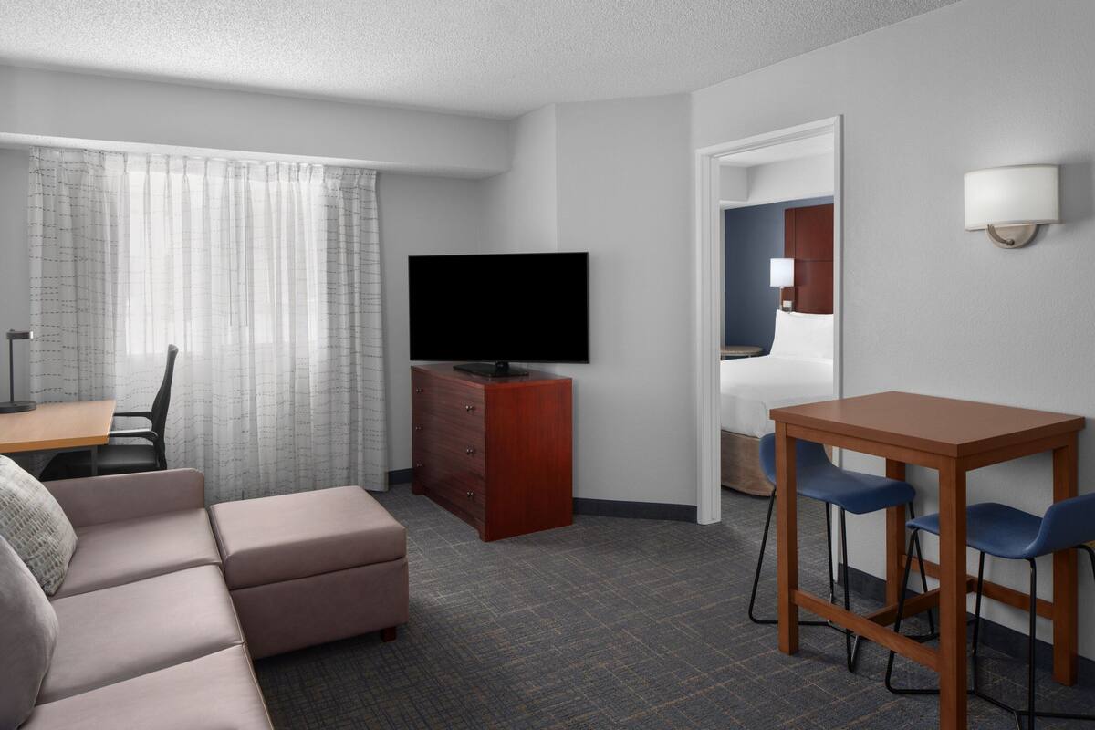 Hotelkamer met een bank, een televisie, een houten dressoir, een bureau met twee stoelen en een deur naar een badkamer.