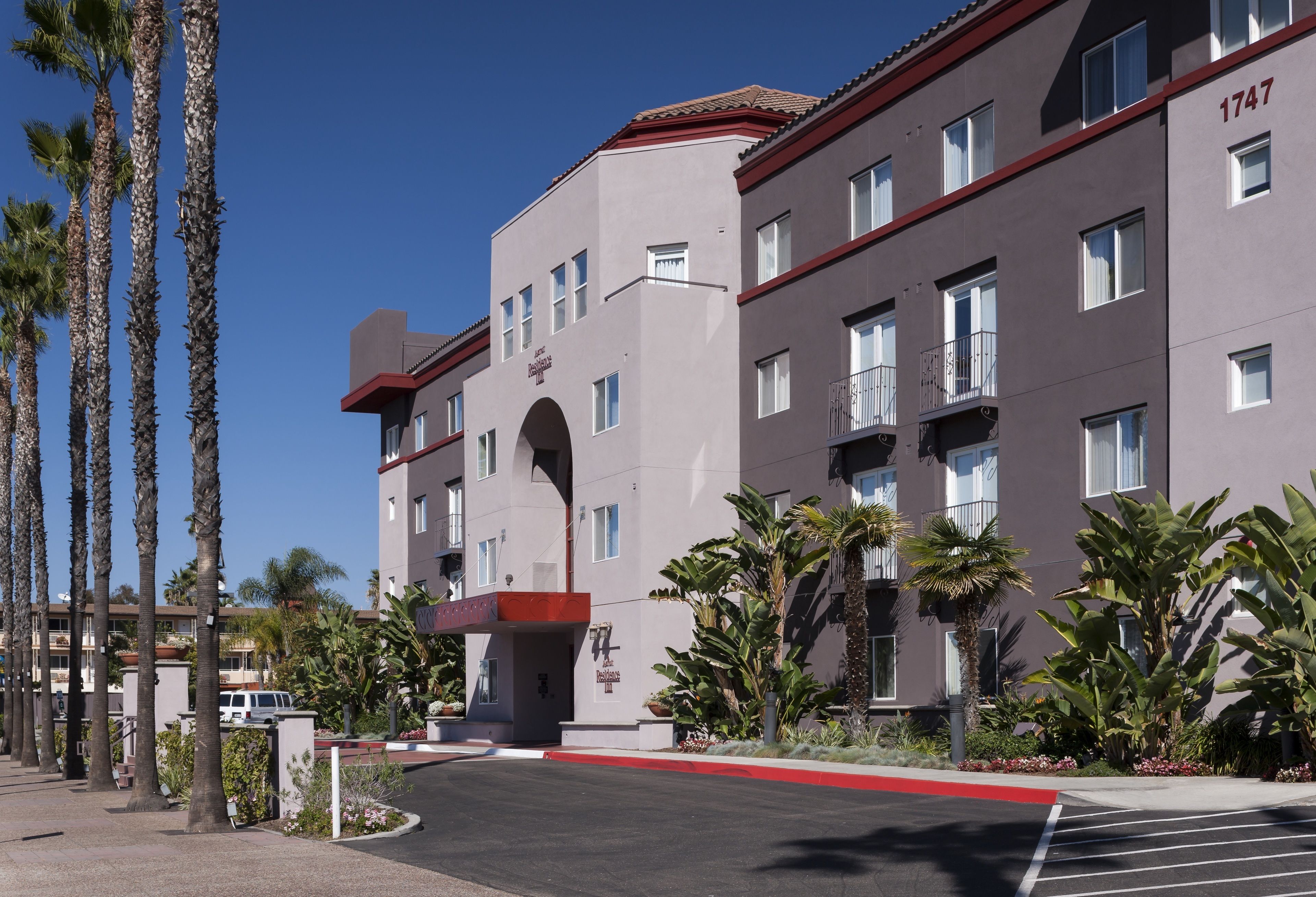 A multi-story hotel with a red and grey facade, numbered 1747, featuring a central archway entrance, surrounded by palm trees and other greenery.