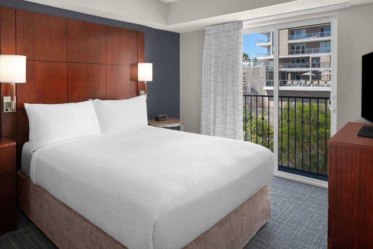 Een hotelkamer met een groot bed, twee bedlampen, een nachtkastje, een televisie en een balkon met uitzicht op de stad.