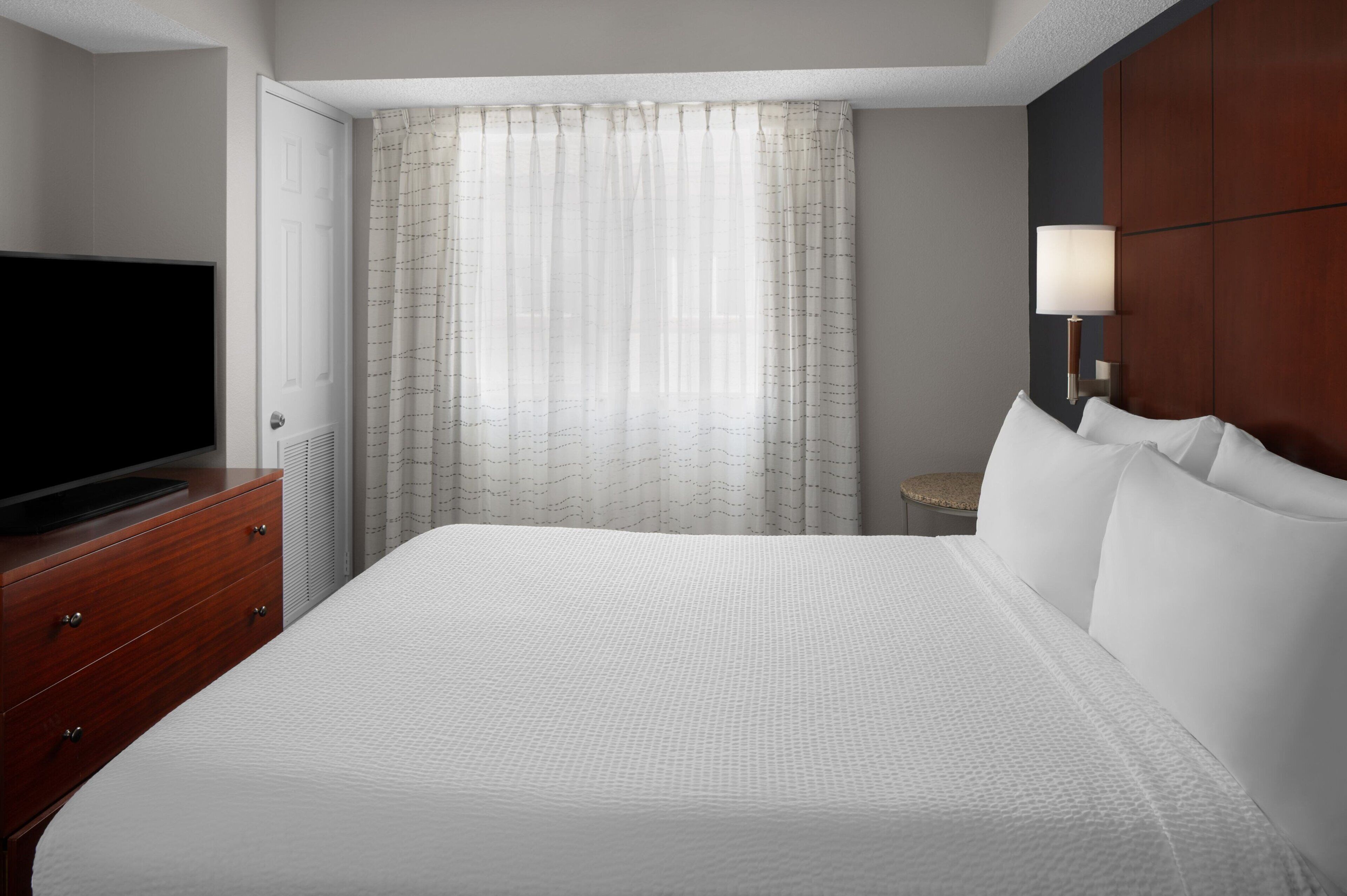 A hotel room with a large bed, a television on a wooden dresser, a lamp, and a window with sheer curtains.