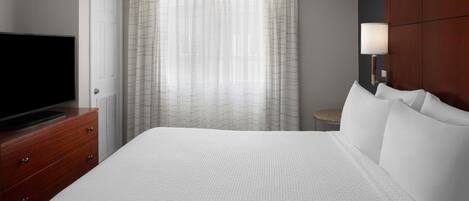 A hotel room with a large bed, a television on a wooden dresser, a lamp, and a window with sheer curtains.