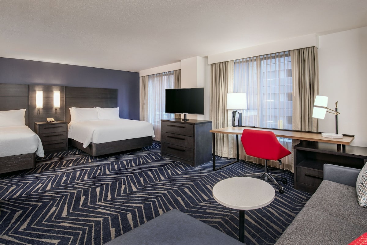 A hotel room with two beds, a desk with a TV, a red chair, and a small round table.