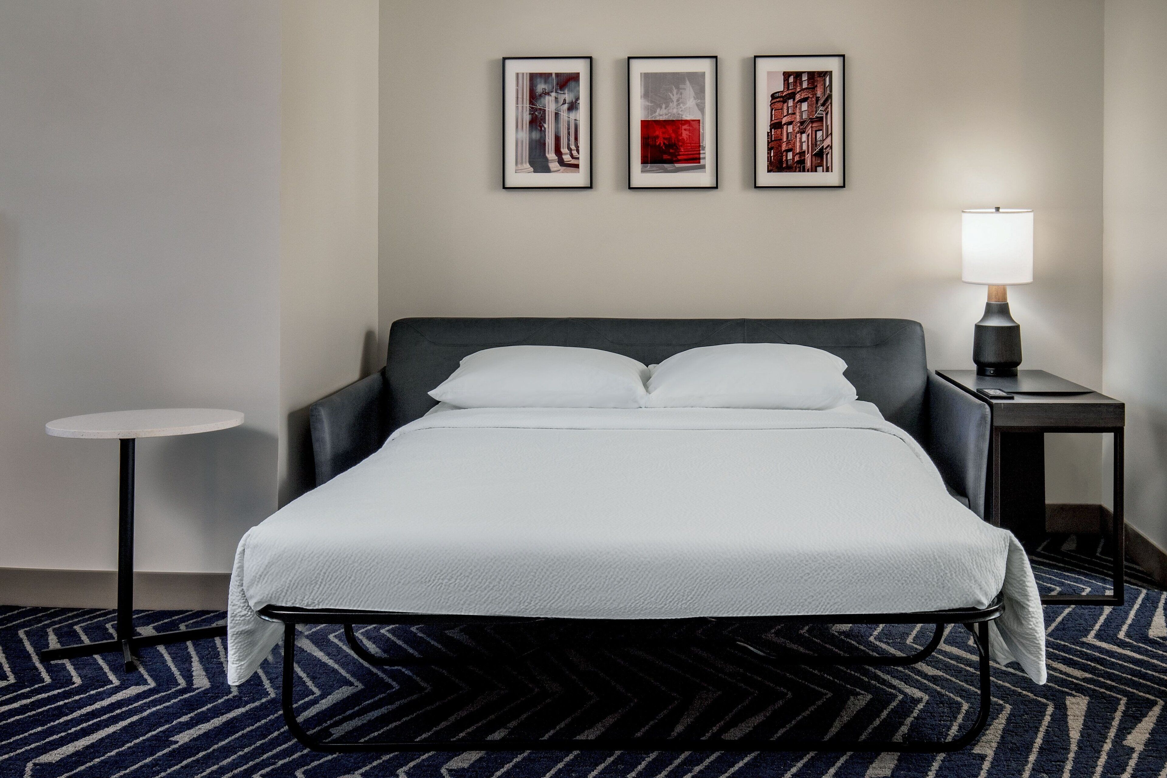 A hotel room with a bed, a small table, a lamp, and three framed pictures on the wall.