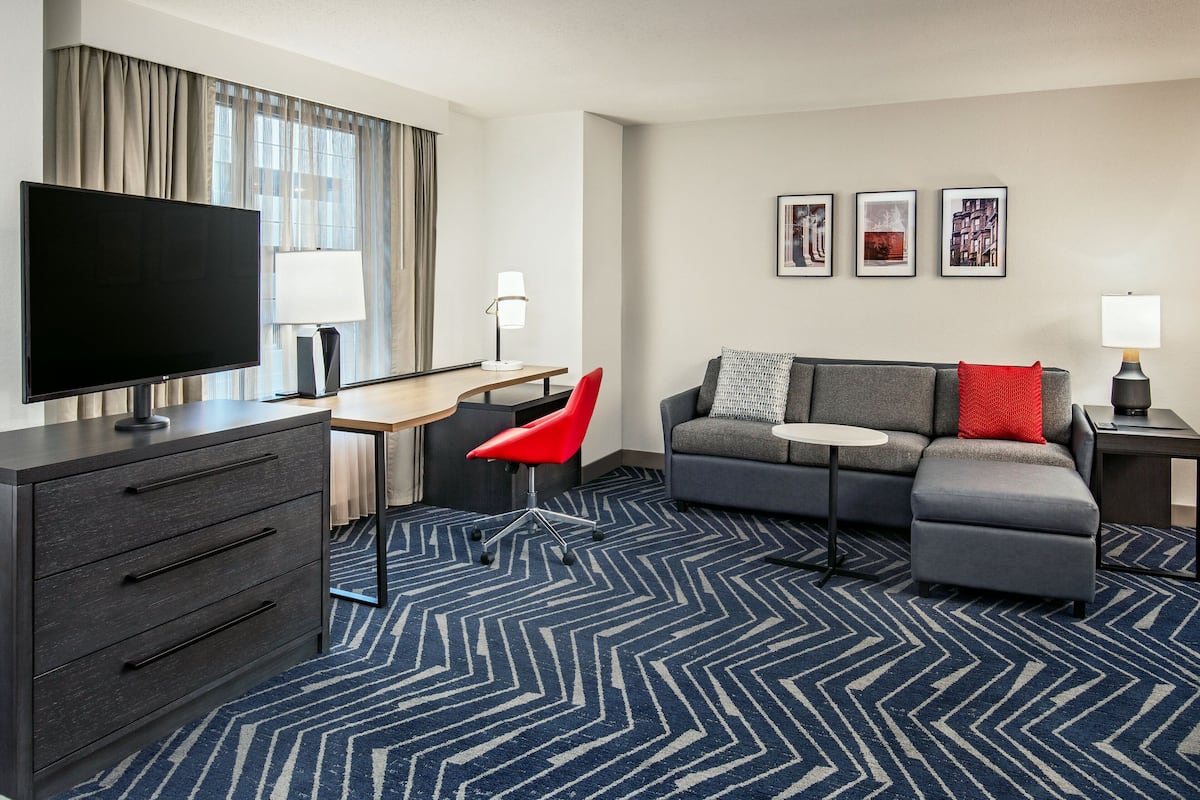 A modern hotel room with a flat-screen TV, a desk, a red chair, a grey sofa, and a patterned carpet.