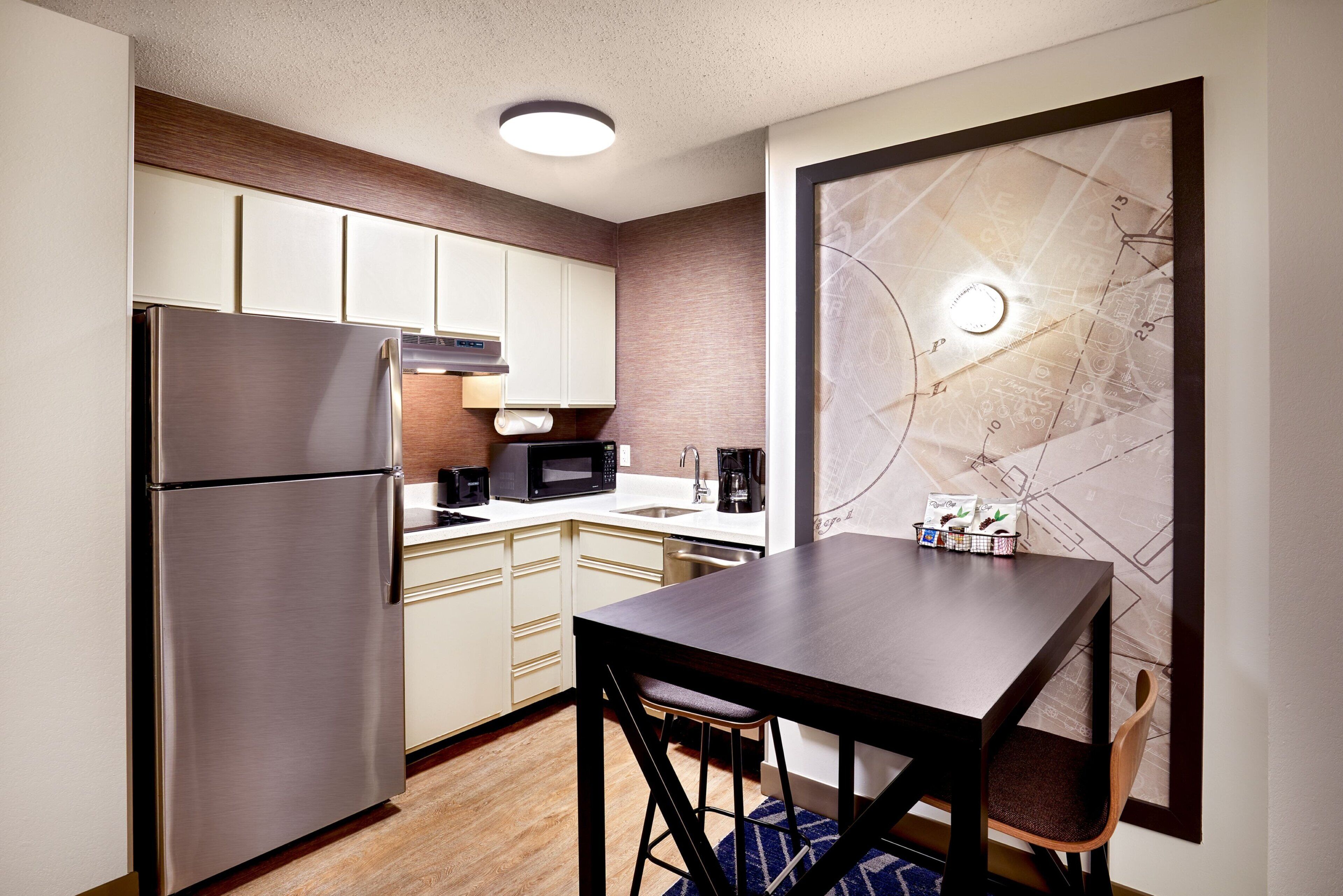 Una cocina compacta con refrigerador, microondas y cafetera. Hay una mesa de comedor con dos sillas y un cuadro con un mapa en la pared.