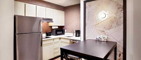 A compact kitchen with a refrigerator, microwave, and coffee maker. There is a dining table with two chairs and a map artwork on the wall.