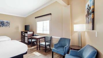 Una habitación de hotel con una cama, un escritorio con silla, un televisor, una ventana con persianas y un cuadro en la pared.