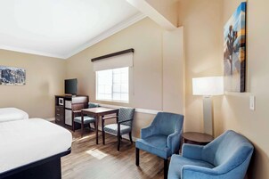 Una habitación de hotel con una cama, un escritorio con silla, un televisor, una ventana con persianas y un cuadro en la pared.