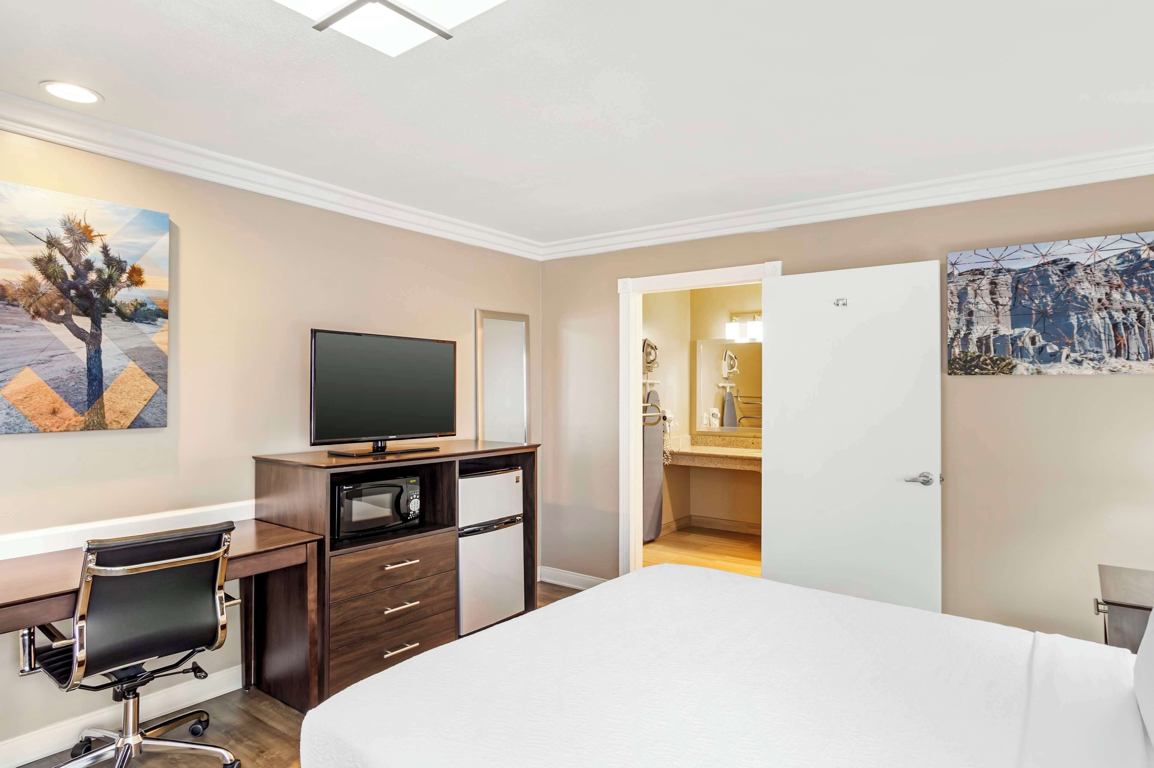 Habitación de hotel con cama, escritorio con silla, televisión, refrigerador pequeño y puerta que da a un baño.