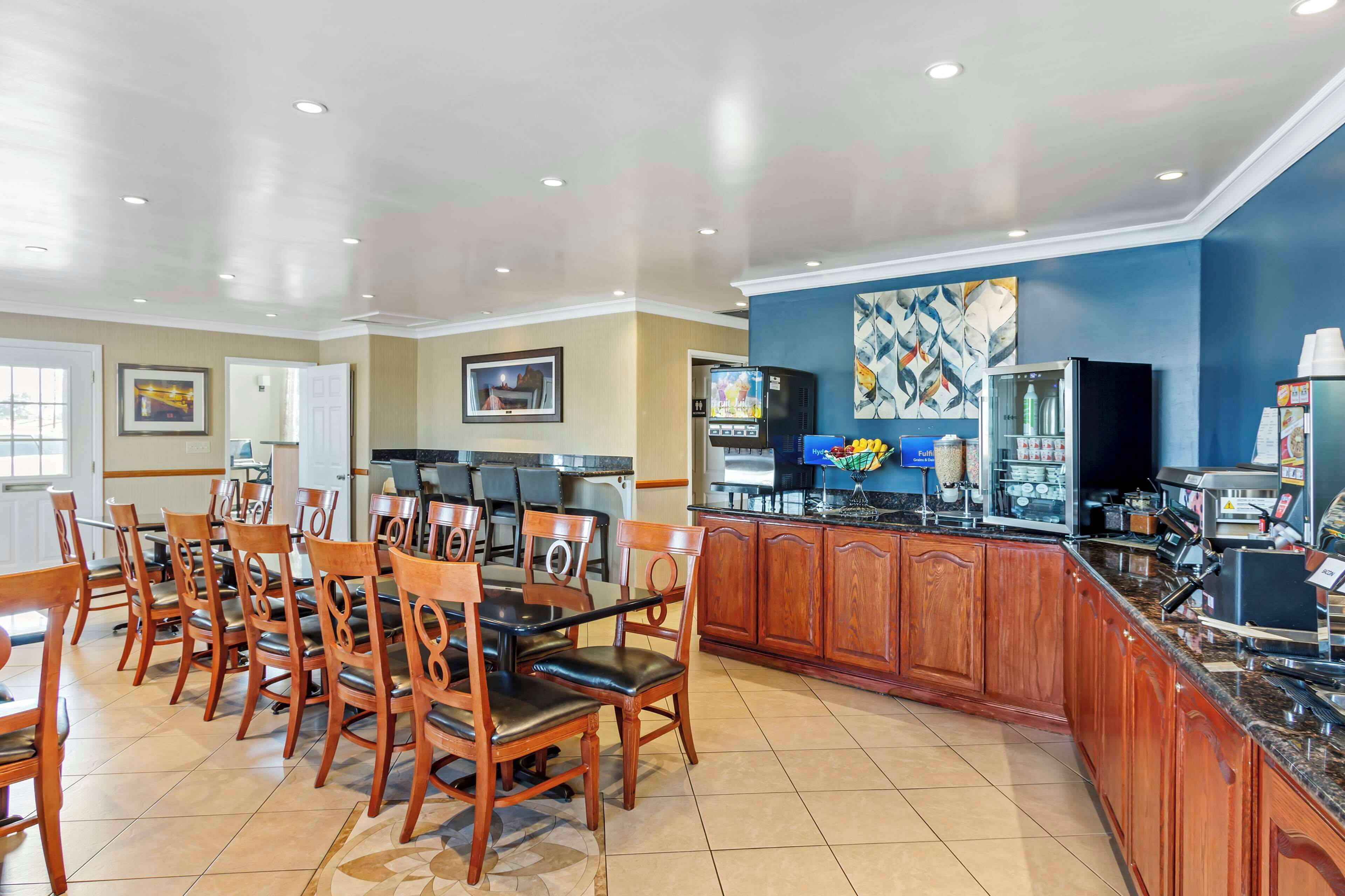 A hotel dining area with wooden chairs and tables, a kitchen counter, and a refrigerator.