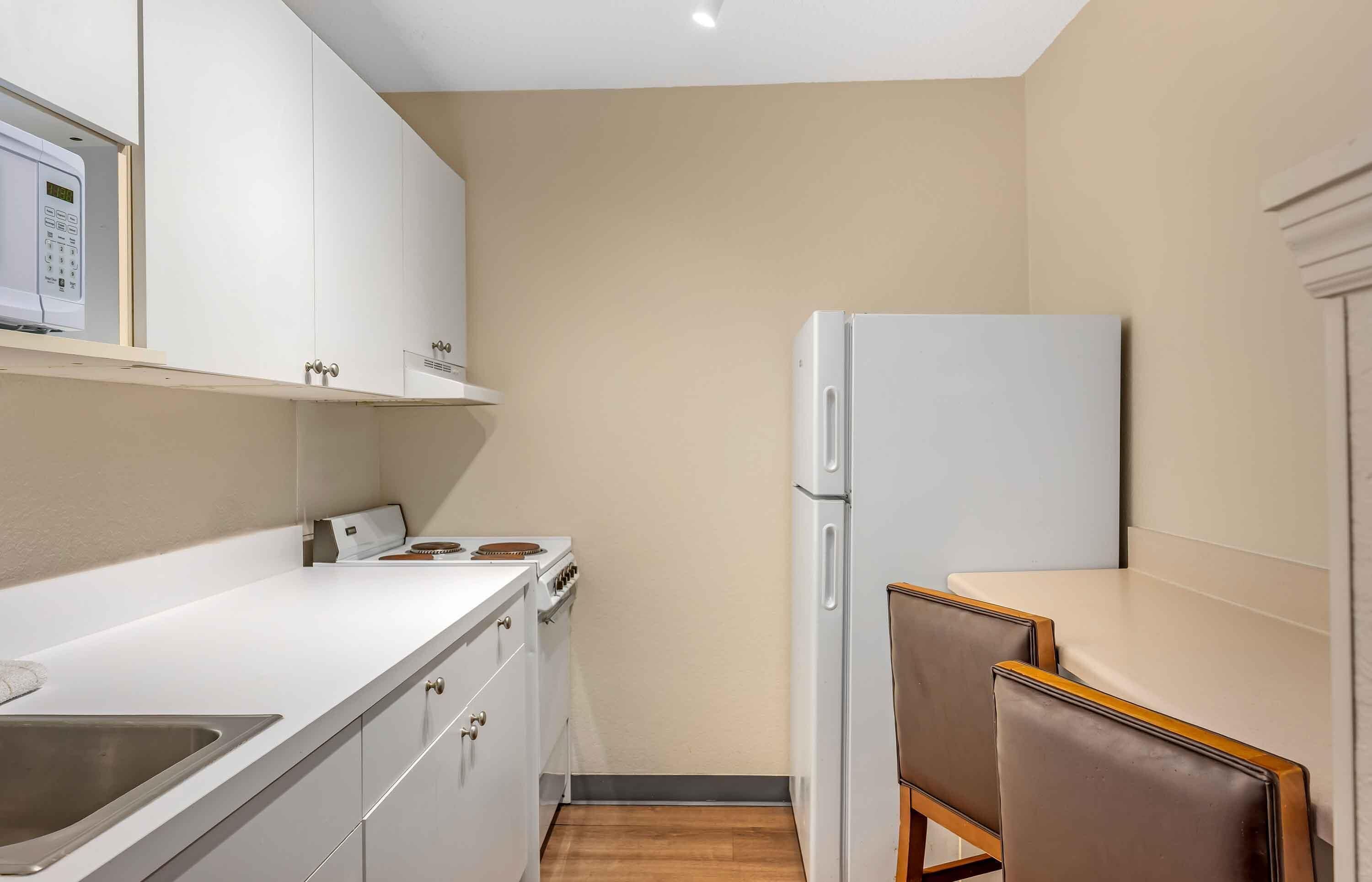 Cocina compacta con gabinetes blancos, microondas integrado, estufa y refrigerador. Hay un pequeño comedor con mesa y sillas.