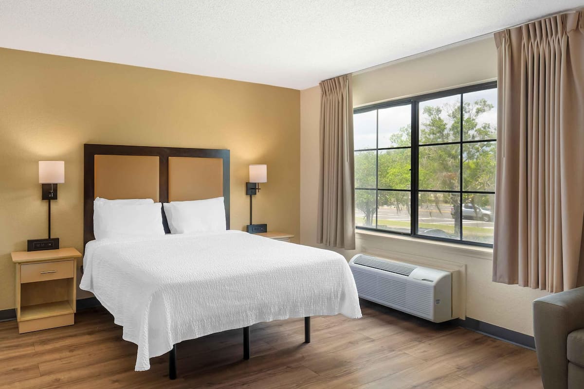 A hotel room with a bed, bedside lamps, a nightstand, a window with curtains, and an air conditioning unit.