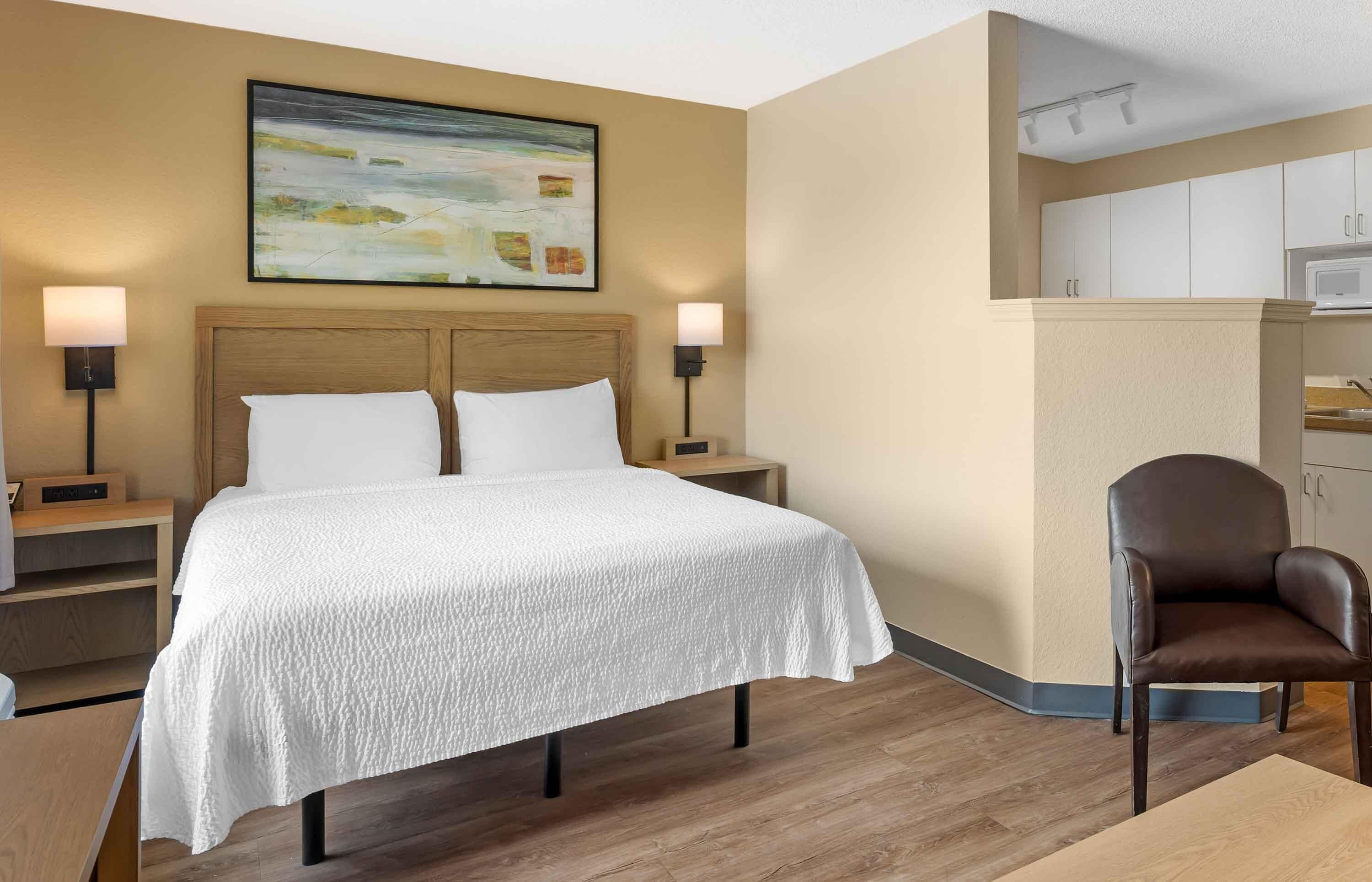 A hotel room with a bed, bedside lamps, a chair, a microwave, and a painting on the wall.