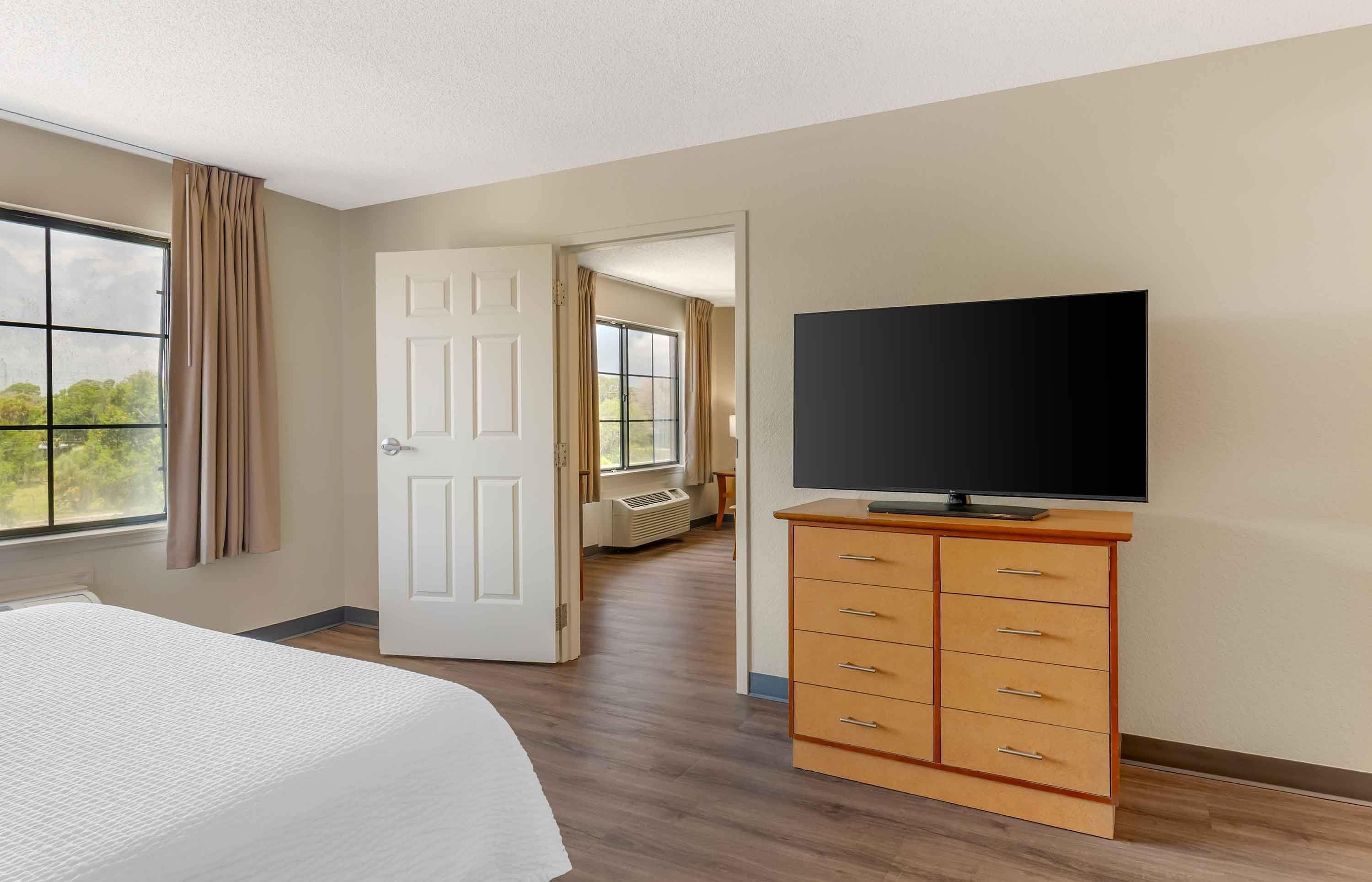 Una habitación de hotel con una cama, un televisor sobre un aparador de madera, una puerta que da a otra habitación y una ventana con cortinas.