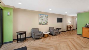Eine Hotelhalle mit Fischgrätparkett, zwei grauen Sessel, einem kleinen runden Tisch, einem Gemälde an der Wand und einer Kommode mit Gegenständen darauf.