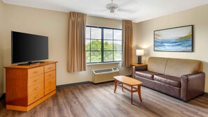 Ein Hotelzimmer mit Flachbildfernseher, einem Holzkoffer, einem braunen Ledersofa, einem kleinen Holztisch, einer Lampe und einem Fenster mit Vorhängen.