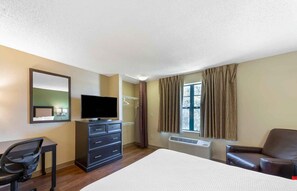 Ein Hotelzimmer mit Bett, Schreibtisch mit Stuhl, Kommode, Fernseher, Fenster mit Vorhängen und Heizkörper.