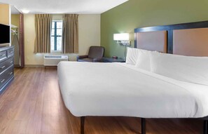 A hotel room with a large bed, a chair, a desk with a lamp, a window with curtains, and a wall-mounted air conditioner. - Extended Stay America Suites Los Angeles Torrance Del Amo Ci (Torrance)