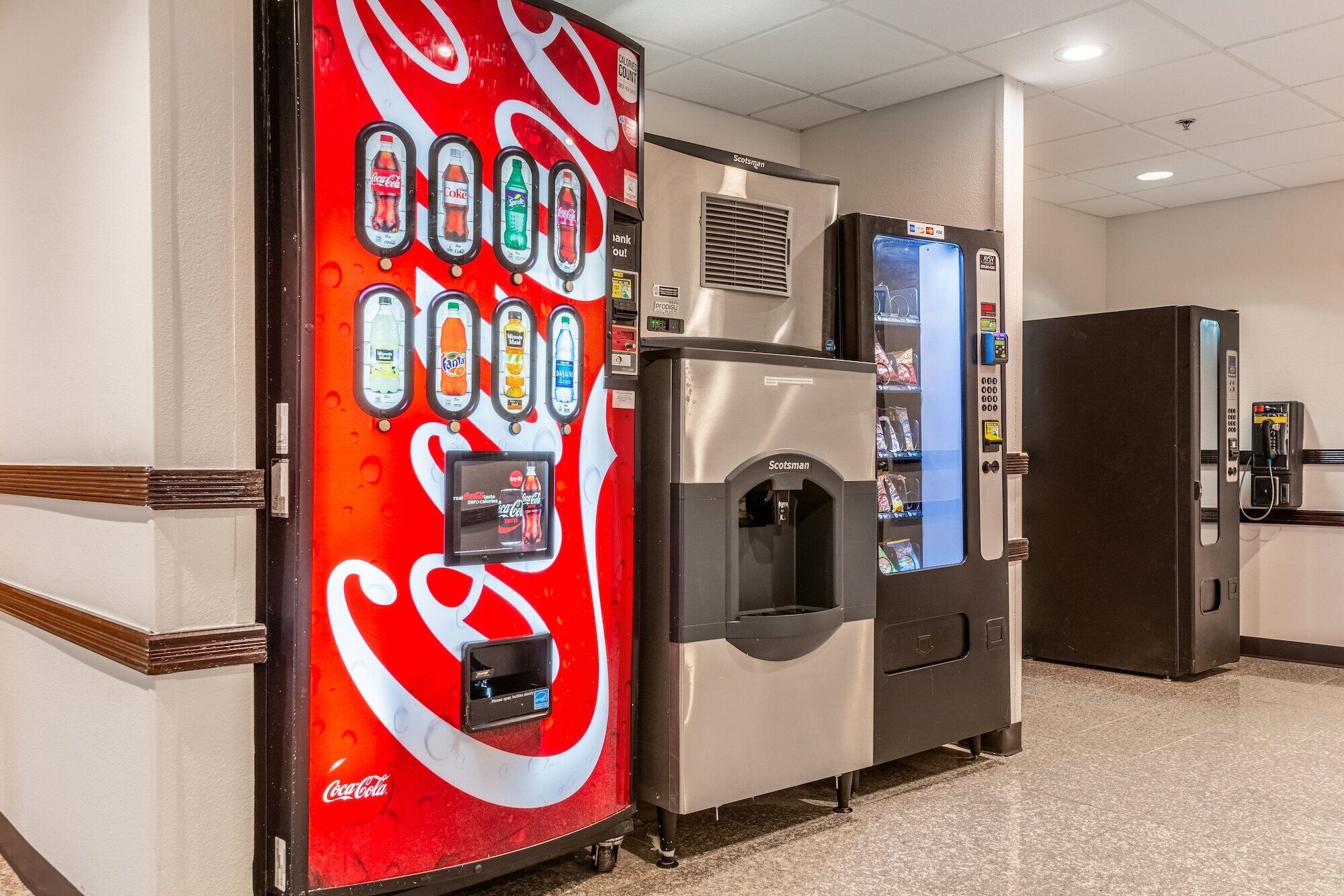 Máquina expendedora con logotipo de Coca-Cola, dispensador de agua y vitrina refrigerada en un pasillo de hotel.