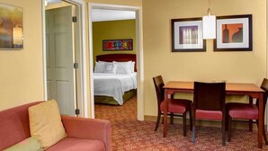 Une chambre d’hôtel avec un canapé rouge, une table à manger en bois et quatre chaises, un lit avec une literie blanche et un tableau coloré accroché au mur.