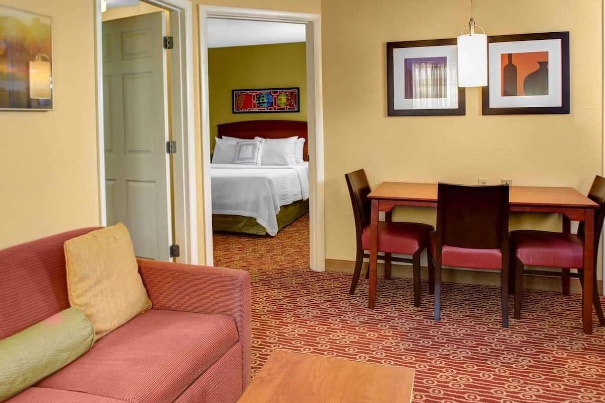 Een hotelkamer met een rode bank, een houten eettafel met vier stoelen, een bed met wit beddengoed en een kleurrijk schilderij aan de muur.