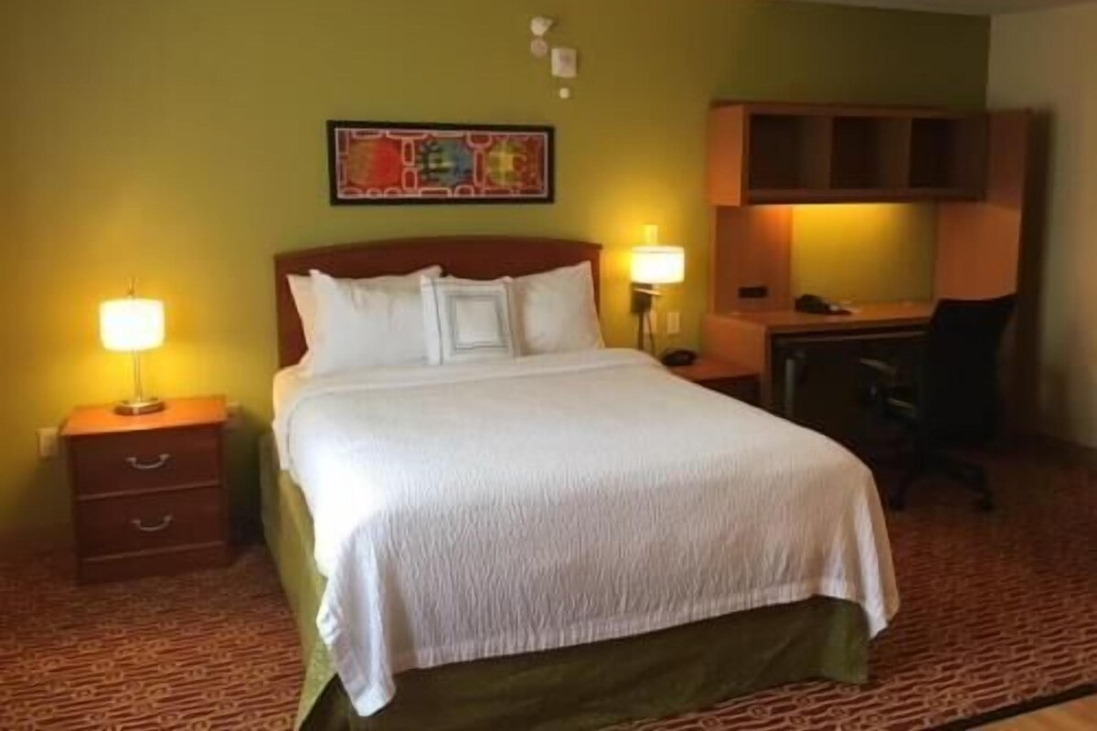 Une chambre d’hôtel comprenant un lit, deux tables de chevet avec des lampes, un bureau avec une chaise et une étagère fixée au mur.
