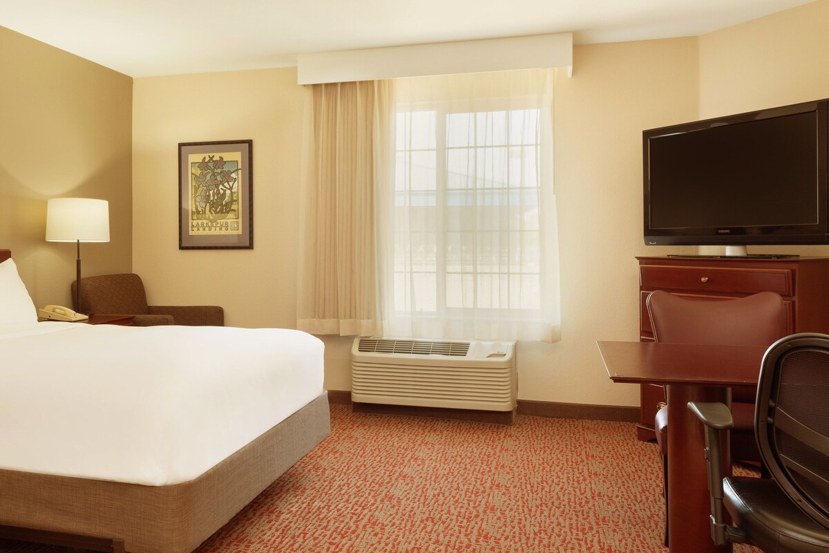 A hotel room with a large bed, a desk with a chair, a television, a window with curtains, and an air conditioning unit.