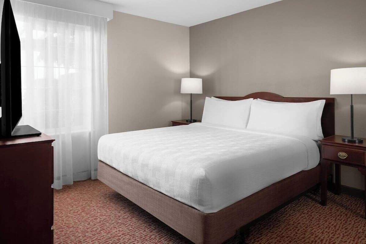 A hotel room with a large bed, two bedside lamps, a wooden nightstand, a TV on a stand, and sheer curtains.