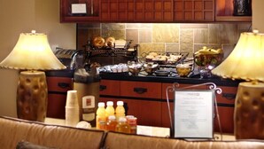 A hotel breakfast area with a counter displaying bread, pastries, and a coffee station. There are two lamps on either side of the counter, and a menu sign is visible.