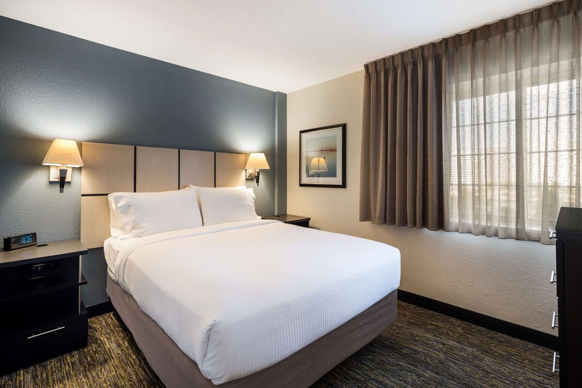 Une chambre d’hôtel avec un grand lit, deux lampes de chevet, une table de chevet, une horloge et une fenêtre avec des rideaux.