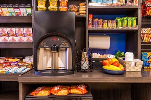 Una estación de café autoservicio con una cafetera, un bol de fruta y diversas opciones de bocadillos y bebidas en estantes.