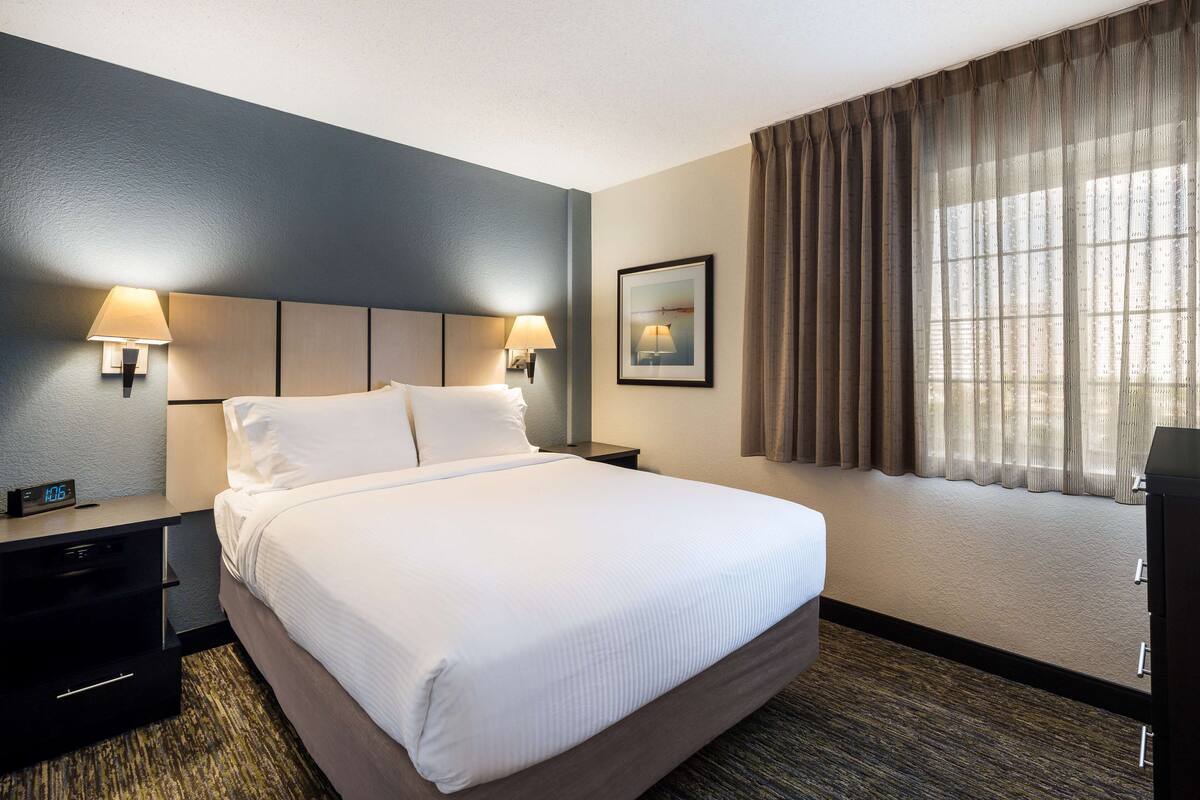 A hotel room with a large bed, two bedside lamps, a nightstand, a clock, and a window with curtains.