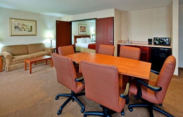 A hotel room with a wooden dining table, four brown chairs, a beige sofa, a small wooden coffee table, a lamp, a framed picture on the wall, and a kitchenette with a microwave.