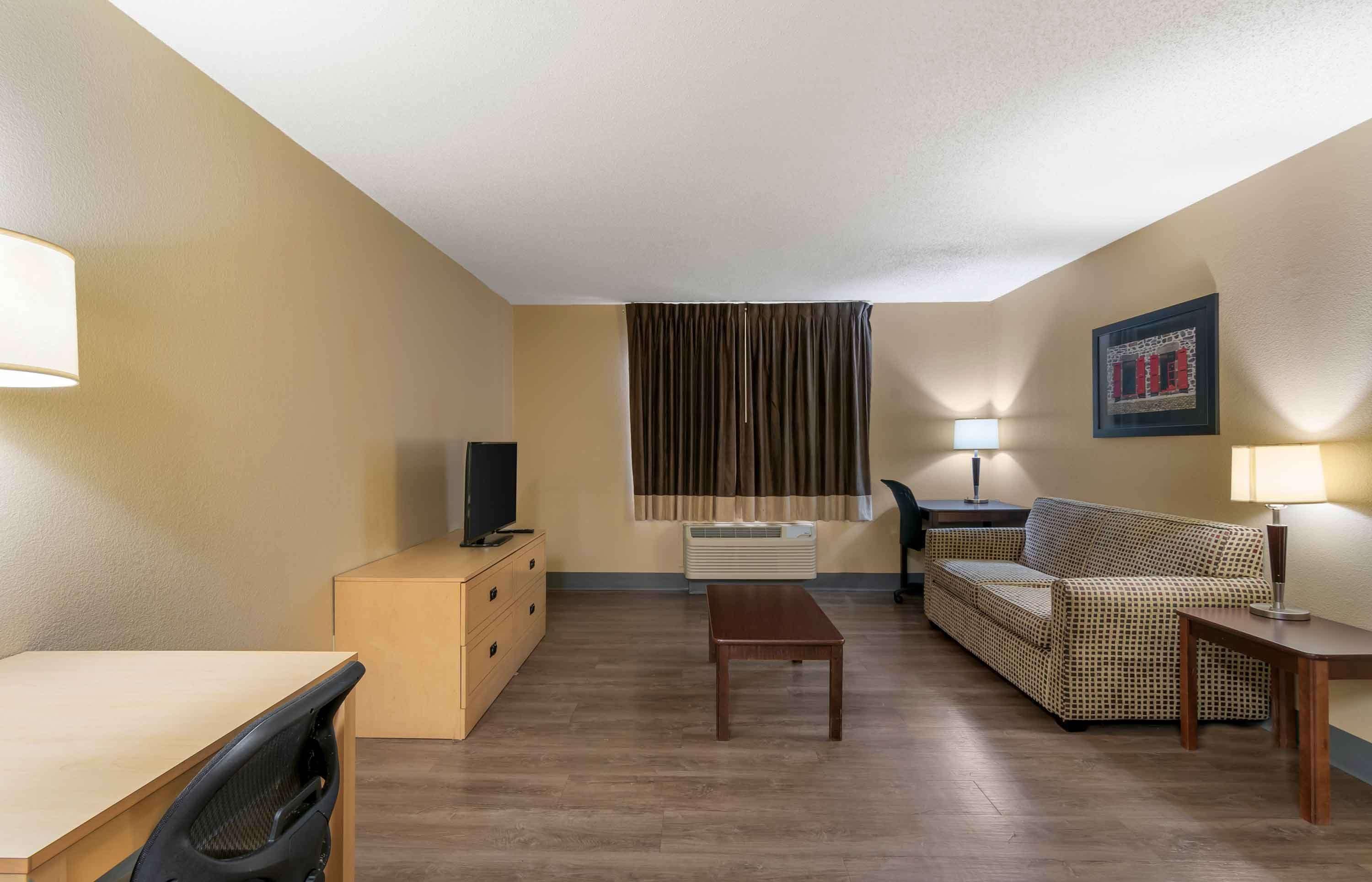 A compact hotel room with a wooden floor, a sofa, a small table, a TV on a dresser, and a window with curtains.