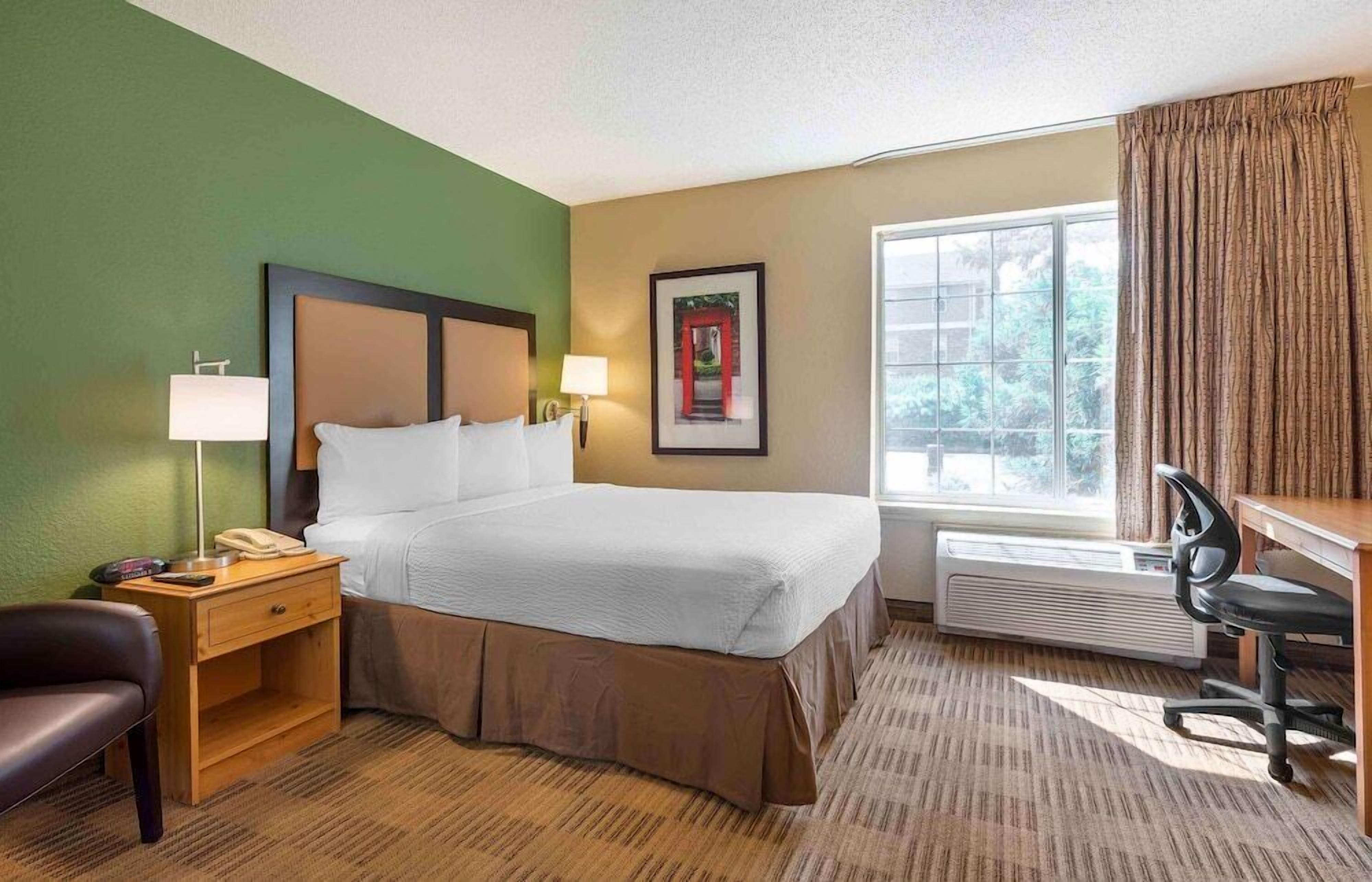 A hotel room with a bed, bedside lamps, a desk, a chair, a framed picture on the wall, and a window with curtains.