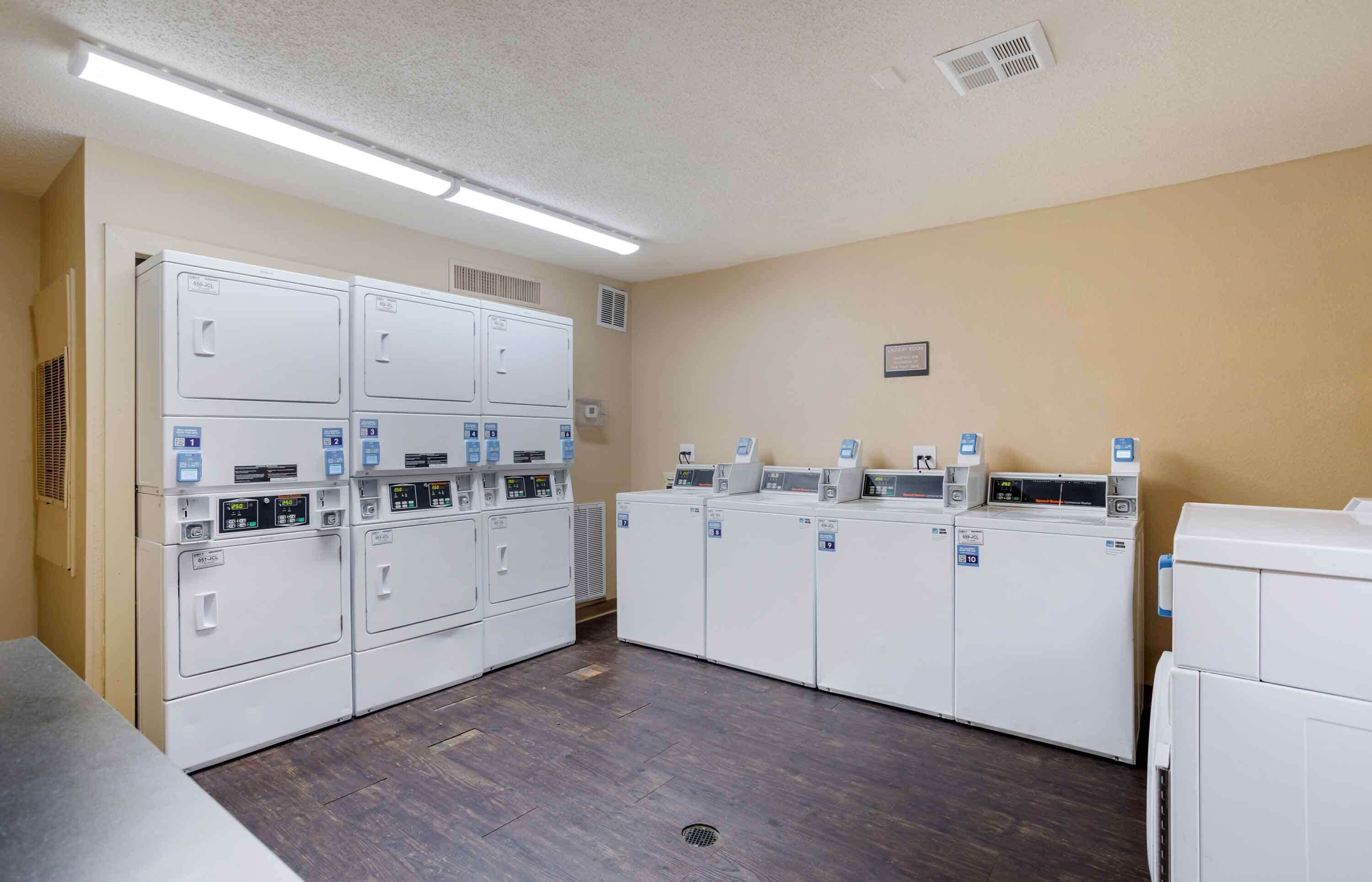 A laundry room with multiple washing machines and dryers, a vent on the wall, and a light fixture above.