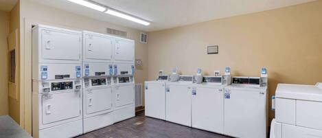 A laundry room with multiple washing machines and dryers, a vent on the wall, and a light fixture above.